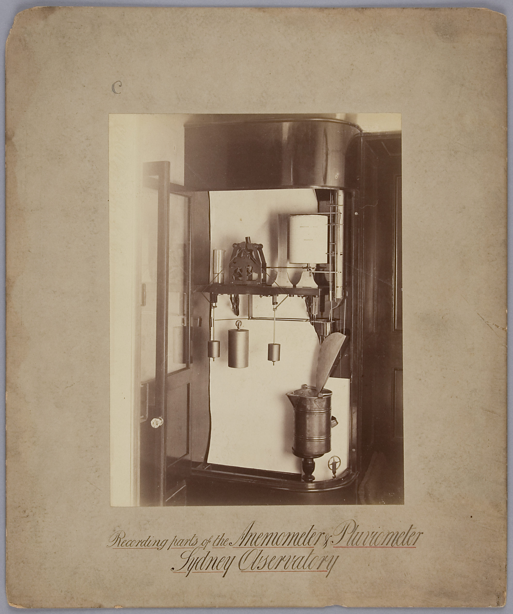 Sepia-toned photograph mounted on an aged piece of paper. The photograph depicts complex measuring machine, approximately as tall as a person, with various weights, pulleys, a water-collecting container and a rolled measurement chart.